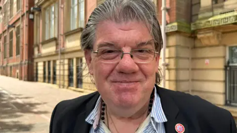 BBC An image of Ged Mirfin outside County Hall in Preston. He is wearing a blue and white striped shirt and black blazer with a Fire Authority pin badge. He is wearing glasses and smiling.