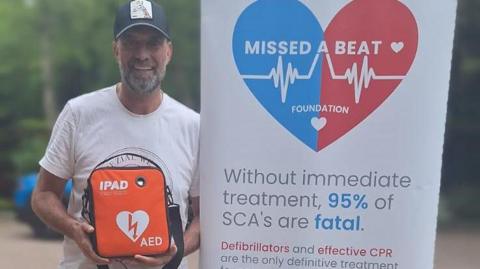 Jurgen Klopp holds a defibrillator next to a banner for Missed a Beat Foundation