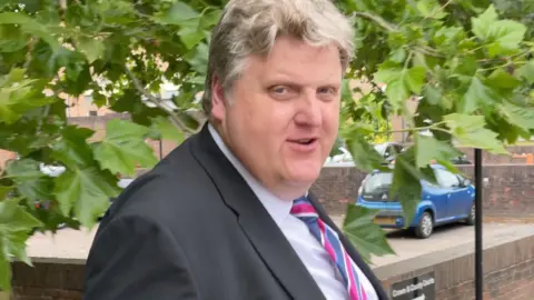 Robby West/BBC Gregory Hill, a man in a black suit jacket, white shirt and striped tie. He has blonde hair and is pictured outside court in Norwich. A tree, a wall and a blue Citroen car are behind him.