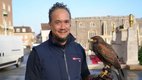 Mikael ChuFoon, a falconer who is looking directly at the camera and smiling. He is wearing a black fleece and navy gilet. He is standing on the top of Norwich marketplace with a Harris's hawk on his arm.