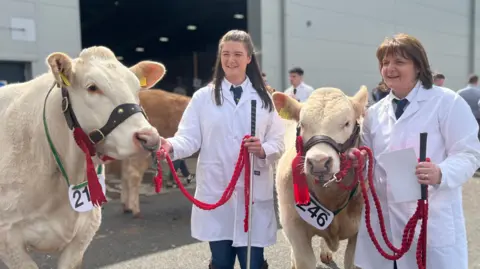 Two woman are standing next to cream and brown cows. They are holding onto red rope which is connected to the cows' harnesses. One woman has long brown hair and the other has short lighter brown hair. Both are wearing a white shirt, white jacket and dark coloured tie. In the picture you can see that one woman is wearing blue denim jeans. They are standing outside a shed at the show.