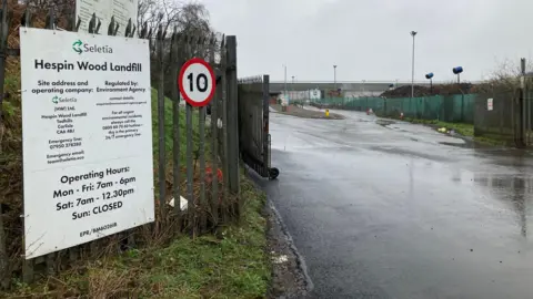 On the left there's a metal fence with a white sign attached which details the opening times for the landfill. On the right there's a tarmac road with floodlights on the right, alongside a green fence.