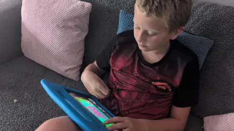 A child sits on a grey fabric sofa using a blue tablet device, with textured cushions in pink and blue placed around them.