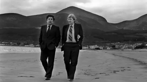 Brendan Murphy A black and white image of two men walking across the beach. One has dark short hair and the other lighter hair. Both are wearing a suit with tie and shirt. The Mourne mountains are pictured in the background, and the waves are crashing on the shore behind them.