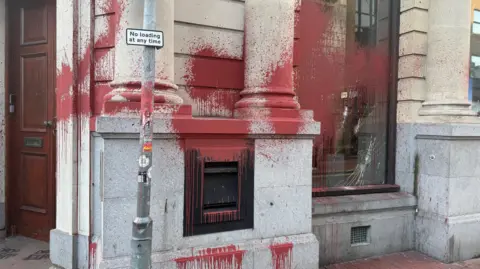 Eddie Mitchell The outside of a bank building with a closed wooden door seen to one side and large pillars either side of high windows. There is a metal deposit box in one wall, and a red substance which looks like paint on the window, which is cracked, and splashed on the walls.