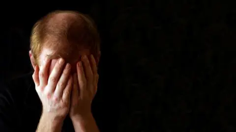 A man with short, light brown hair holds his head, which is bowed forwards, covering his face.