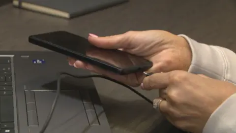 A close-up of female hands plugging a USBC cable into a mobile phone, held over the keyboard of a laptop.
