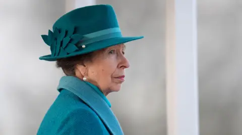 Princess Anne, wearing pearl earrings, a teal tweed coat, shirt, and hat with a flower detail on, looks into the distance. There is a grey blurred background behind her and a white pillar.