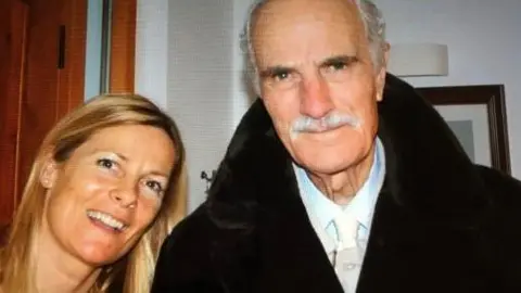 The picture shows two people standing indoors. One person is wearing a dark coat with a wide collar over a light-coloured shirt. In the background, there is a wooden door, a white wall, a framed picture, and a wall-mounted light fixture. 