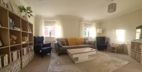 An example of another crisis house run by Alabare - it is a living room with two royal blue arm chairs, a grey sofa with a wooden table on a beige patterned rug with a large book case displaying books and plants and a TV.