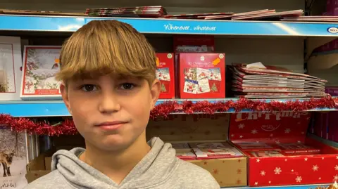 BBC Ethan standing in front of a Christmas shelf in Kinnerley Shop. There are Christmas cards and tinsel behind him. 