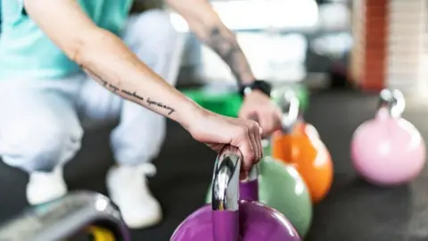 A person leans while squatting on a floor, their hands leaning on the metal handles of kettle bells. The bells are different colours. The person has a tattoo of words on their right arm.