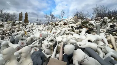 A mound of mannequins, from full bodies, to legs and heads and torsos. The pile goes as far as the eye can see. Body parts are white, black and cream.