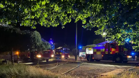 Essex County Fire and Rescue Service Three fire engines pictured in the dark with their headlights on. There are hoses running to them over muddy ground. There are trees in the foreground.