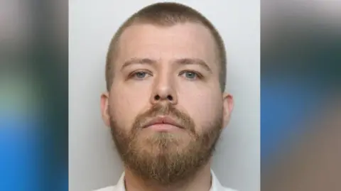 Derbyshire Police Mugshot of a man in a white shirt. He has a buzzcut and a light brown beard.