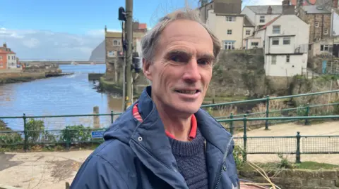 Colin Harrison dressed in a blue waterproof jacket and a navy blue woolly jumper stands in front of the small fishing village of Staithes on the edge of the edge of the North Yorkshire Moors. 