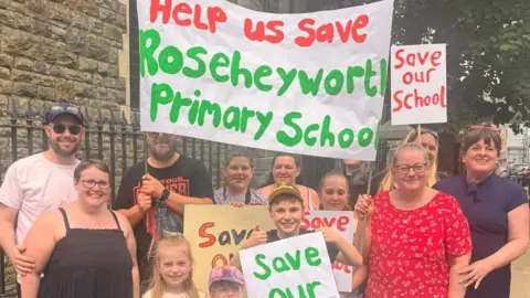 A group of adults and children holding signs saying Save our School and a big banner which says 'Help us save Roseheyworth Primary School'.