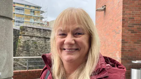 A woman with blonde hair and a purple coat stands in front of the Derry Walls
