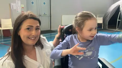 In a sports hall with a blue floor a woman in a white top smiles to the camera as she has her hand on the back of her daughters wheelchair. The woman, who is kneeling beside her daughter is on the left of her daughter. Her daughter looks of to the right of the camera as she sits wearing a lilac top.