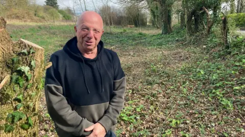 LDRS A man, wearing a black and green hoodie, with a bald head and glasses, stands in front of a clearing lined with trees near Curry Rivel on the Somerset Levels.