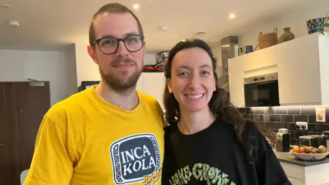 A man in a yellow top stands next to a woman in a black one. He has short brown hair and glasses. She has long dark brown hair
