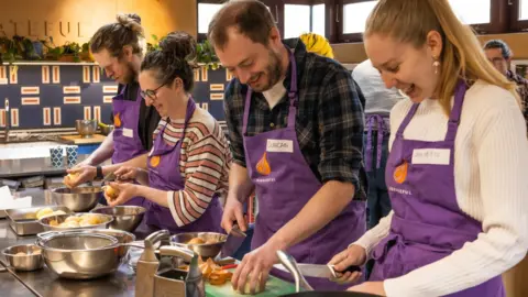 Fede Rivas A group of men and women are standing in a kitchen wearing a purple apron with name tags. There are two men and two women working on a table with kitchen utensils. They are all chopping, peeling and preparing food. They are all smiling while keeping an eye on what they are doing. Two people, a man and woman, are holding big sharp knives. The other man and woman are peeling potatoes. 