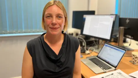 Dr Laura Goodfellow pictured at her desk in her office, with her laptop behind her. She has bobbed blonde hair and is wearing a black short-sleeved top.