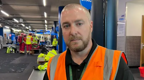 Lily-May Symonds/BBC A close-up image of Dave King wearing an orange high-visibility vest indoors, with emergency responders and equipment visible behind them in a ski facility setting.
