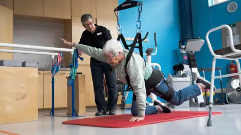 Shaftesbury Icanho A woman supported by a ceiling harness poses on a yoga mat on all fours, with her right arm and left leg raised and outstretched, watched by a woman in dark clothing whose name badge on her chest in not readable.