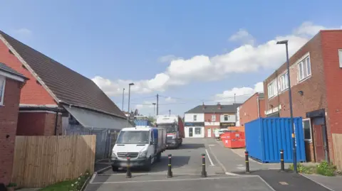 A road with bollards in the foreground and buildings wither side. There are two lorries parked on the left.