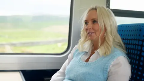 Nick Wooley/BBC A smiling blond lady with tan skin wearing a white long-sleeved shirt and blue sweater vest