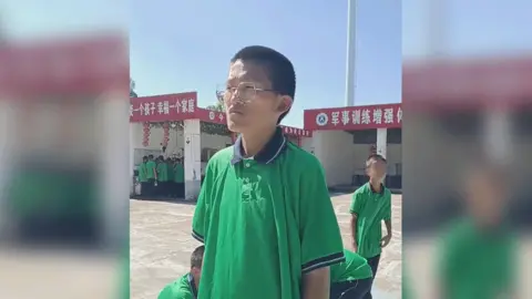 An image taken from video footage sent to Enxu's parents, taken of Enxu in an open concrete area inside Shengbo school. Her hair is cut short and she is wearing a green polo shirt with navy blue and white trim, which looks too big for her. She is standing straight and looking into the distance to the left of the camera.