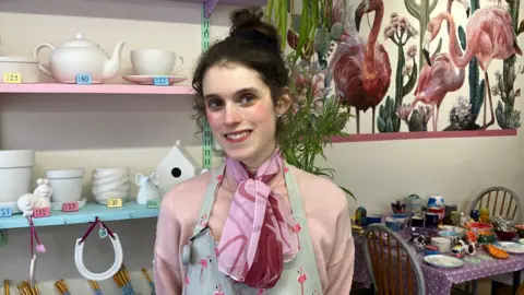 A young woman with dark brown hair tied in a high bun is smiling at the camera She is wearing a pink jumper, a small pink scarf and an apron with pink flamingos printed on it.