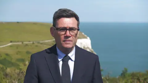 Mid shot of Andy Burnham wearing a black suit and black tie with black-framed glasses. In the distance is the white cliffs of Dover.