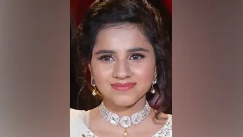 A smiling woman with tied back black hair wearing gold earrings and a diamond looking necklace and white dress 