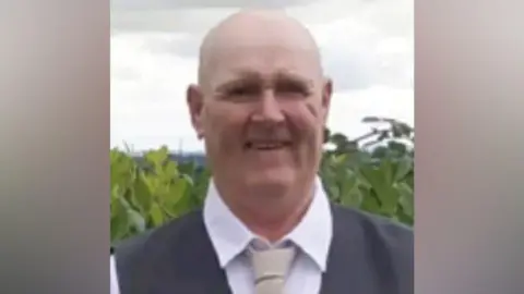 A bald man smiling at the camera wearing a white shirt, black waistcoat and cream tie.