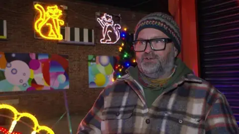 Tom Ingall/BBC Simon Thirkill wearing a checked shirt and a green scarf and multicoloured hat, stood in front of some of the lights. 