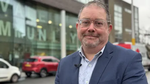 A man with short grey hair, a grey beard, wearing glasses, a blue suit jacket and a light blue shirt, smiles while he stands next to a road.