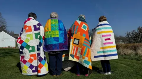 BMQG Four people standing on grass with their backs to camera and quilts draped over their shoulders