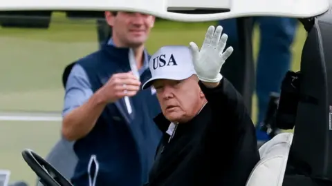 Reuters Donald Trump waving from a golf buggy, wearing a white "USA" cap.