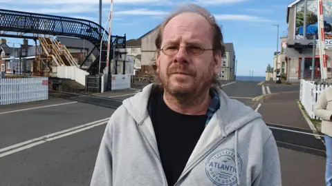 A man, with balding brown hair and brown stubble, looking at the camera. He is wearing glasses, a black stop and a grey zip-up hoodie. He is standing at a crossroad and there is a black bridge visible to his left. 