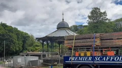 Scaffolding being erected at the fenced-off bandstand. A lorry with building materials is parked in front of it. The structure has ornamented railings and a grey roof with a cupola on top of it. It is surrounded by trees.