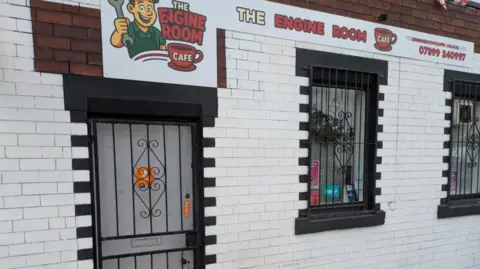The front of a cafe, signposted as 'The Engine Room Cafe'. It is a white brick building with a locked gate over the front door.