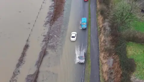 Eric Bird A white car, a silver car and a blue van driving in flood water. 