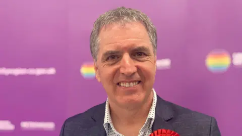 A man with short grey hair smiles at the camera. He is wearing a white shirt with small triangles on and a grey jacket. He's standing against a purple backdrop and you can see a red rosette on his jacket, as he's a Labour politician. 