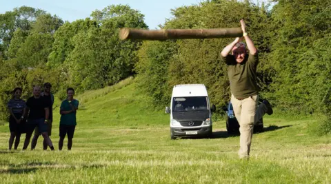 PA A man wearing khaki trousers and a green long-sleeved top. He is throwing a long wooden pole ahead of him using both hands, while several onlookers watch behind him. They are competing on a large grassy field, with a sloped hill and trees in the background.