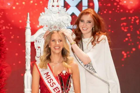 Alan Strutt/Miss England Grace Richardson wearing a red dress being presented with a tiara by a woman wearing white