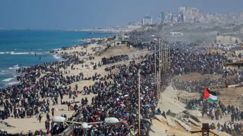 Rekaman drone Reuters menunjukkan massa orang berjalan di sepanjang jalan pesisir untuk kembali ke utara Gaza, sebuah kota di depan mereka dan laut di sisi kiri mereka, dengan bendera Palestina terlihat. 