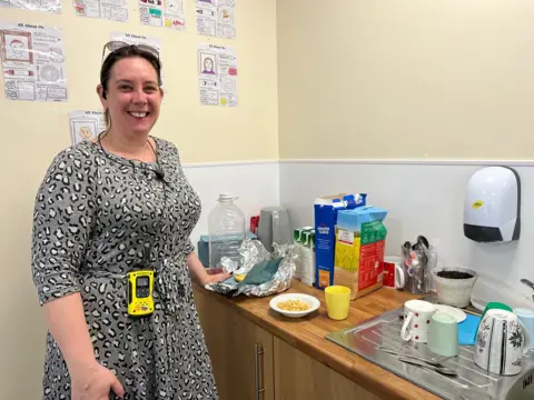 A woman wearing a grey animal print dress with glasses on her head. She is in front of a small kitchen  that has a metal sink draining board with mugs on, and cereal boxes, milk cartons and some other utensils on. She also has a small yellow and black gadget dangling hanging from a string lanyard around her neck.