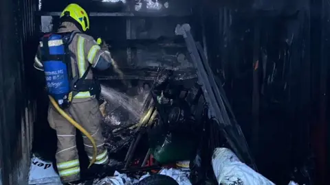 Firefighter at the scene of the blaze in Cornwall. The firefighter is wearing fire safety equipment. The firefighter is stood inside of the burnt container and is spraying water to dampen down hotspots. Items within the container are black and burnt.